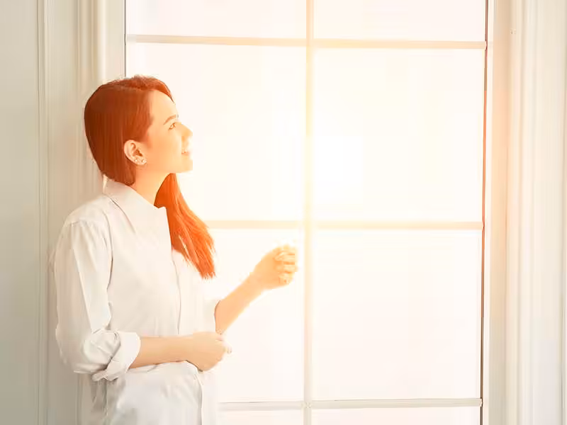 Young beautiful woman standing against window watching at sunris